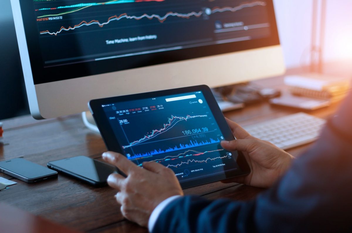 Empresario revisando el mercado de valores en tableta digital y un ordenador de sobremesa con un gráfico de bolsa en pantalla